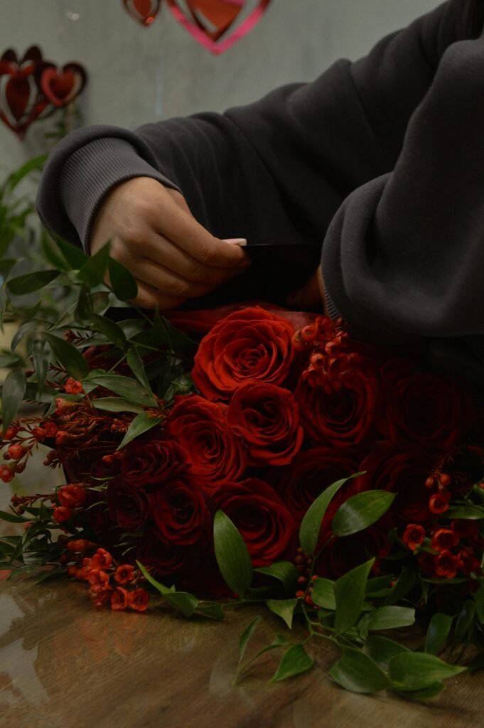 A florist arranges a stunning red rose bouquet with green leaves and berries, indoors.
