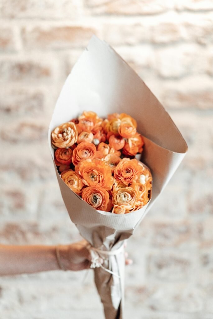 A bright bouquet of orange ranunculus flowers wrapped in paper, held against a textured brick wall.
